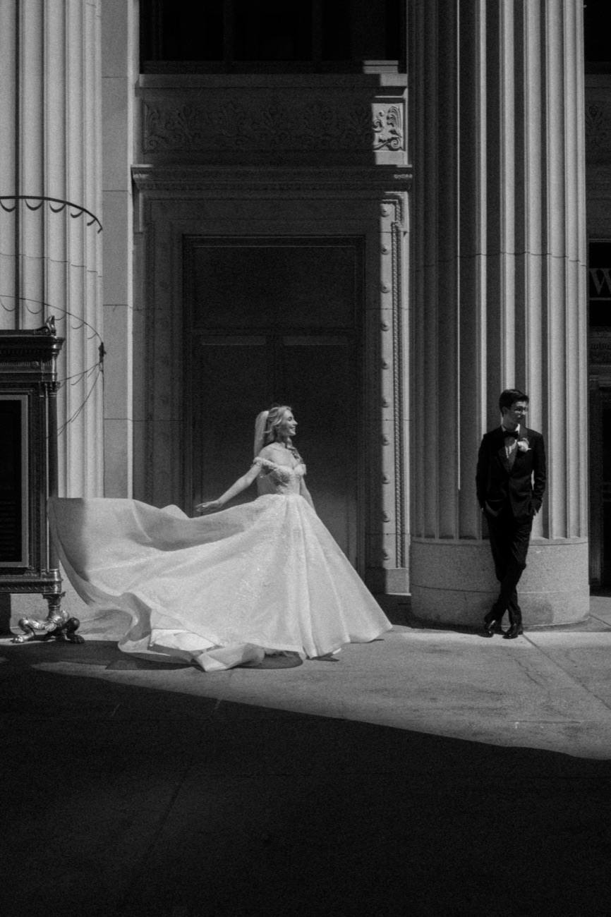 A black and white view of a bride's gown caught mid-motion beside a colonnade