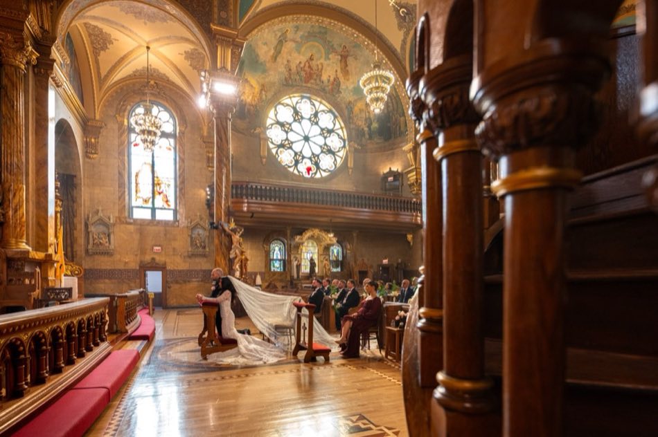 A bride and groom kneeling together in prayer beneath a rose window