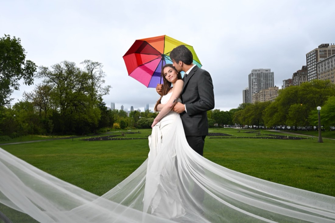 A couple sheltering under a rainbow umbrella with the Chicago skyline behind them